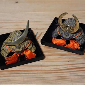 以下が含まれることがあります： 黒い正方形のフレームに収められた2つのミニチュア日本の侍の兜。1つは大きく湾曲した銀色の角、もう1つは金色の角があります。どちらもオレンジ色の房と複雑なディテールが特徴です。