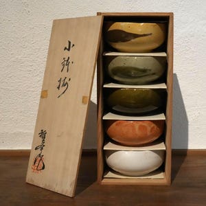 以下が含まれることがあります： 木製の箱に蓋があり、黄色、緑、オレンジ、白など様々な色の陶器のボウルが5つ入っています。箱の蓋には日本語の文字が書かれています。