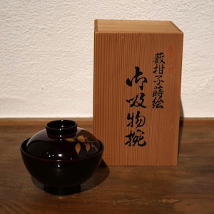 以下が含まれることがあります： 葉の模様が入った、蓋付きの濃い茶色の漆塗りの日本のスープボウル。その隣には、日本語の文字が書かれた木箱があります。ボウルは木製の表面にあります。