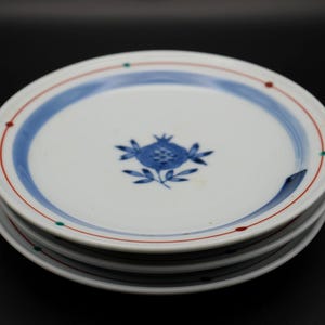 May include: Four white ceramic plates stacked on top of each other. Each plate has a blue pomegranate design, a blue border, and a red and green dotted rim. The plates are displayed against a black background.
