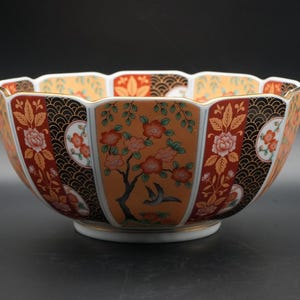 Puede incluir: Un cuenco decorativo de cerámica con borde festoneado. El cuenco presenta un diseño floral y de pájaros en naranja, rojo, negro y dorado. El interior es de color naranja claro y la base es blanca.