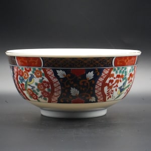 Puede incluir: Un cuenco de cerámica decorativo con interior blanco y diseño exterior detallado. El cuenco presenta una base color crema, una banda de patrones rojos y negros y un diseño central con flores y pájaros. El borde está adornado con oro.