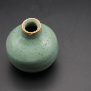 Puede incluir: Un jarrón redondo de cerámica de color celadón con una pequeña abertura y un borde ligeramente ensanchado. El jarrón tiene un cuerpo liso y redondeado y una textura moteada. El borde es de color marrón claro. Es un objeto decorativo.