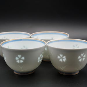 Puede incluir: Cuatro cuencos de cerámica blanca con un diseño floral. Cada cuenco tiene un borde azul y un patrón floral azul claro. Los cuencos están dispuestos sobre una superficie negra.