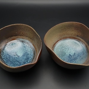 以下が含まれることがあります： ユニークなデザインの手作りのセラミックボウル2つ。ボウルは茶色の外側と青とターコイズの内側を持っています。ボウルは有機的な形をしており、食べ物を提供したり、装飾品として使用したりする可能性があります。