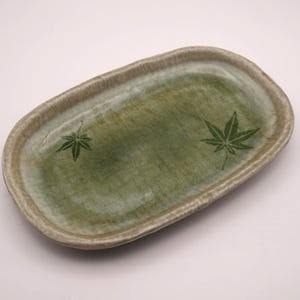 以下が含まれることがあります： 緑色の内側と薄茶色の縁が付いた、楕円形のセラミック製サービング皿。濃い緑色のカエデの葉の模様が2つ、緑色の表面に印刷されています。この皿はおそらく食べ物の提供に使用されます。