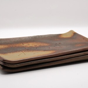May include: Three rectangular ceramic plates stacked on top of each other. The plates are brown and tan, with a slightly curved edge. The plates have a rustic, handmade appearance, suitable for serving food.