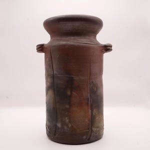 Puede incluir: Un jarrón de cerámica hecho a mano con un cuerpo cilíndrico y una parte superior ensanchada. El jarrón presenta una rica paleta de colores terrosos de marrones, rojos y toques de negro. Dos pequeñas asas están en los lados. El jarrón tiene un aspecto rústico y texturizado.