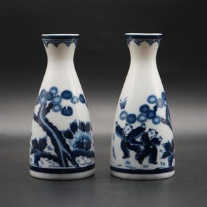 Puede incluir: Dos botellas de sake de cerámica blanca con diseños florales y figurativos azules. Las botellas tienen un cuello estrecho y una base más ancha. Una botella presenta un diseño de árbol y flores, la otra figuras.