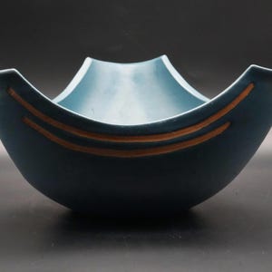 May include: A handcrafted, blue ceramic bowl with a unique, curved design. The bowl features two parallel, horizontal, orange-brown lines on the exterior. The bowl's interior is a lighter shade of blue, creating a striking contrast.