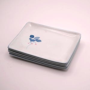 May include: A stack of four square white ceramic plates. Each plate has a blue floral design and a blue border. The plates are stacked on top of each other, with the top plate showing the full design.