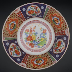 Puede incluir: Un plato de cerámica decorativo con un diseño floral. El plato presenta un centro blanco con un colorido arreglo floral. El borde exterior tiene un patrón repetitivo de formas geométricas y motivos florales rojos, azules y verdes.