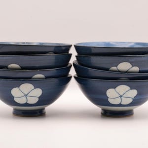 以下が含まれることがあります： 紺色のセラミックボウル8個セット。各ボウルは白い花の模様と青色の内側が特徴です。ボウルは4つずつ2つのグループに積み重ねられており、その形状とデザインを示しています。