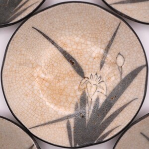 May include: A set of ceramic plates with a cream-colored, crackled glaze. Each plate features a black and gray floral design, including a stylized flower and long leaves. The plates have a dark rim.
