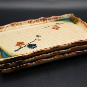 Puede incluir: Una pila de cuatro platos rectangulares de cerámica. Cada plato tiene un fondo beige con un diseño floral en naranja y negro. Los bordes son de color marrón oscuro con una textura ondulada.