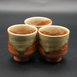 May include: Three handmade ceramic cups with a rustic design. The cups feature a combination of earthy tones, including brown, green, and orange. The interior of the cups is a light beige color.