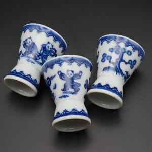 以下が含まれることがあります： 青い手描きのデザインが施された、白い磁器製の卵カップが3つ。各カップには、人物や木など、異なるシーンが描かれています。縁と底は青い縁で装飾されています。