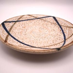 以下が含まれることがあります： クリーム色のベースと茶色とネイビーブルーの抽象的な線が入った、斑点のあるセラミックプレート。 プレートはわずかに盛り上がった縁と素朴で手作りの外観をしています。