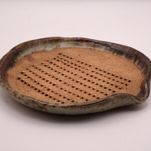 Puede incluir: Jabonera de cerámica hecha a mano con interior marrón y borde verde y marrón. El plato tiene una serie de pequeños agujeros cuadrados en el centro para el drenaje. El plato tiene forma ovalada con un borde ligeramente elevado.