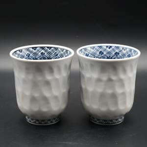 以下が含まれることがあります： 外側がテクスチャ加工され、内側が青と白の模様の白いセラミックティーカップ2つ。各カップの底には青色の幾何学模様があります。カップは暗い背景に表示されています。