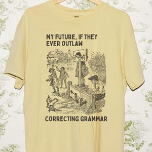 Peut inclure: T-shirt jaune clair avec un motif noir et le texte "MY FUTURE, IF THEY EVER OUTLAW CORRECTING GRAMMAR". Le motif est une illustration vintage de personnes sur une place de ville. Le t-shirt est suspendu à un cintre en bois.