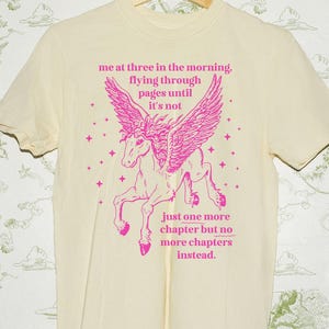 May include: Cream-colored t-shirt with a pink winged horse graphic and text. The text reads "me at three in the morning, flying through pages until it's not. Just one more chapter but no more chapters instead."