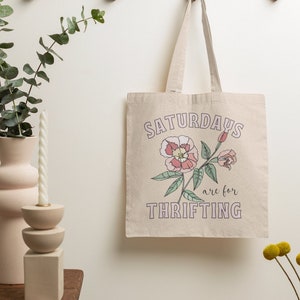 May include: A beige canvas tote bag with the text "Saturdays are for thrifting" in pink and green lettering. A pink flower with green leaves is printed on the bag.