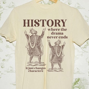May include: Cream-colored t-shirt with the word "HISTORY" in brown, along with the phrase "where the drama never ends" and "it just changes characters." Two figures in robes are depicted. The shirt is hanging on a wooden hanger.