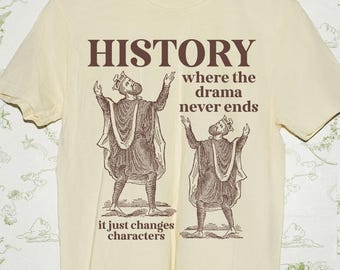 Pode incluir: Camiseta creme com a palavra "HISTORY" em castanho, juntamente com a frase "where the drama never ends" e "it just changes characters". Duas figuras em trajes são retratadas. A camiseta está pendurada em um cabide de madeira.