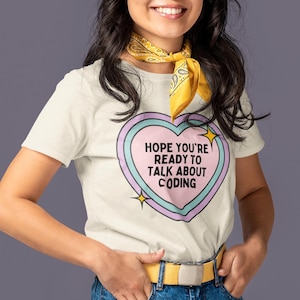 May include: A cream-colored t-shirt with a heart-shaped graphic that reads "HOPE YOU'RE READY TO TALK ABOUT CODING." The graphic is outlined in pink and blue with yellow stars. The person is wearing blue jeans and a yellow belt.