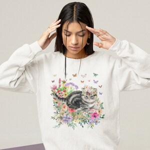 May include: A white sweatshirt with a graphic print of a kitten wearing a flower crown and surrounded by flowers and butterflies.