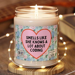 Könnte beinhalten: Eine brennende Kerze in einem Glas mit einem floralen Etikett und einem rosa Herz mit der Aufschrift "Smells Like She Knows A Lot About Coding". Die Kerze steht auf einem runden Holzuntersetzer, mit Lichterketten und einer Gitarre im Hintergrund.