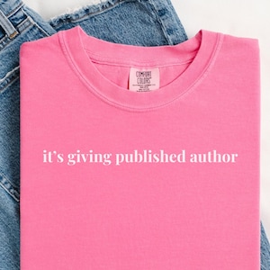 Peut inclure: Un t-shirt rose vif avec le texte blanc "it's giving published author". Le t-shirt est plié sur un jean bleu. Le t-shirt a un col rond et des manches courtes. Le t-shirt est fait d'une matière douce.