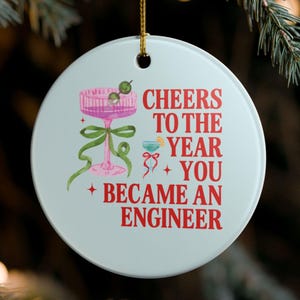 May include: A round white ceramic ornament hanging by a gold string. The design features a pink cocktail glass with a green ribbon and two olives, a small blue drink, and red text reading: CHEERS TO THE YEAR YOU BECAME AN ENGINEER.