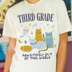 Puede incluir: Una camiseta de manga corta color crema con el texto THIRD GRADE en la parte superior y PAWSITIVELY OUT OF THIS WORLD en la parte inferior. El gráfico incluye varios gatos ilustrados con cascos espaciales flotando entre estrellas y planetas.