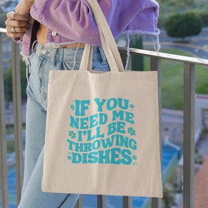 May include: A white canvas tote bag with a blue graphic that says "If you need me I'll be throwing dishes".