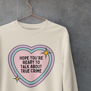 Puede incluir: Una sudadera de color crema con un gráfico de corazón rosa. El corazón tiene un contorno azul y morado y presenta el texto "Espero que estés listo para hablar de crímenes reales".