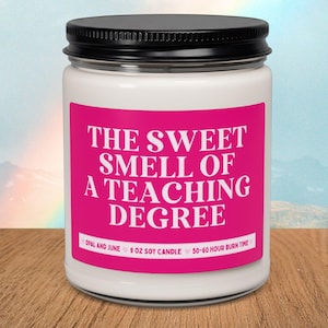 May include: A clear glass candle jar with a black lid and a bright pink label. The label features white text that reads THE SWEET SMELL OF A TEACHING DEGREE, OPAL AND JUNE, 9 OZ SOY CANDLE, and 50-60 HOUR BURN TIME in a clean font.