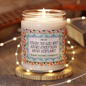 Pode incluir: Uma vela num frasco de vidro transparente com cera branca e um pavio aceso. O rótulo apresenta uma borda xadrez colorida e o texto: SMELLS LIKE YOU'RE THE GIRL WHO KNOWS EVERYTHING ABOUT AIRPLANES, OPAL AND JUNE, 9 OZ SOY CANDLE.
