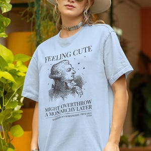 May include: A light blue t-shirt featuring a black graphic print of a historical figure with the text "FEELING CUTE MIGHT OVERTHROW A MONARCHY LATER" and "MAXIMILIEN ROBESPIERRE, PROBABLY" along with small star icons printed on the front chest.