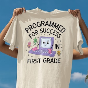 Puede incluir: Camiseta color crema con el texto "PROGRAMADO PARA EL ÉXITO EN PRIMER GRADO". El diseño presenta una computadora de dibujos animados con una cara sonriente, rodeada de ilustraciones de robots. La camiseta se muestra contra un cielo azul.
