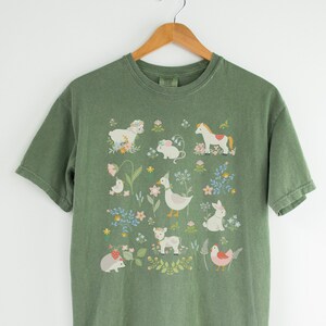 May include: A green t-shirt with a white graphic of various animals and flowers. The animals include a horse, a duck, a rabbit, a cow, a chicken, a hedgehog, a mouse, and a lamb.