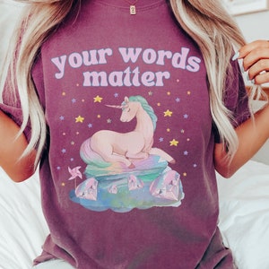 May include: A purple t-shirt with the text "your words matter" in pink and blue letters. A cartoon unicorn with a rainbow mane and tail is sitting on a pile of colorful crystals with stars in the background.