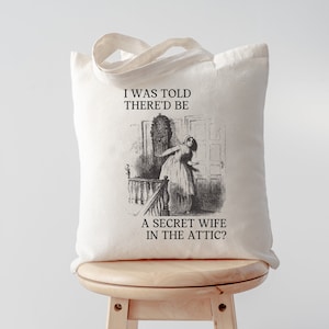 May include: A white canvas tote bag with black text and an illustration of a woman looking up a staircase. The text reads "I was told there'd be a secret wife in the attic?"