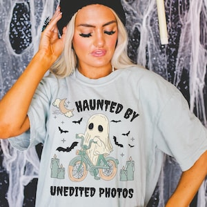 Könnte beinhalten: Ein hellblaues T-Shirt mit einem Aufdruck eines Geistes, der ein Fahrrad fährt. Der Text "Haunted by Unedited Photos" ist auf dem Shirt gedruckt.