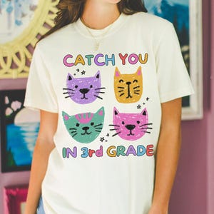 Puede incluir: Camiseta color crema con cuatro caras de gatos de dibujos animados en morado, amarillo, verde y rosa. El texto "CATCH YOU IN 3rd GRADE" está impreso encima y debajo de las caras de los gatos en letras coloridas. La camiseta tiene mangas cortas.