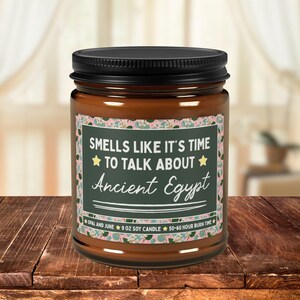May include: A 255g soy candle in a brown glass jar with a black lid. The label features a dark green rectangular centre with the text "SMELLS LIKE IT'S TIME TO TALK ABOUT Ancient Egypt" surrounded by a pink and green floral patterned border.