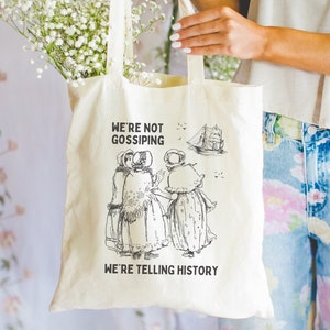 May include: A white canvas tote bag with black text and a black and white illustration of three women in dresses. The text reads "We're not gossiping, we're telling history."