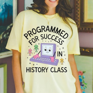 May include: A light yellow t-shirt with the text "PROGRAMMED FOR SUCCESS IN HISTORY CLASS" in a playful font. The graphic features a computer with a face, robot characters, and stars.