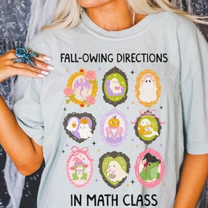 May include: Pale blue t-shirt with the text "FALL-OWING DIRECTIONS IN MATH CLASS." The shirt displays framed illustrations of ghosts, pumpkins, and a frog. The illustrations are in various colours, including pink, green, and purple.